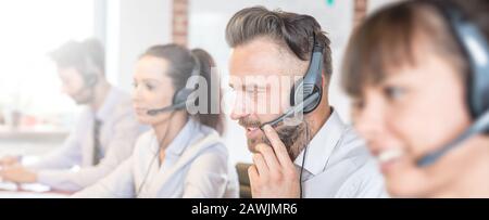 Call center worker accompanied by his team. Smiling customer support operator at work. Young employee working with a headset. Stock Photo