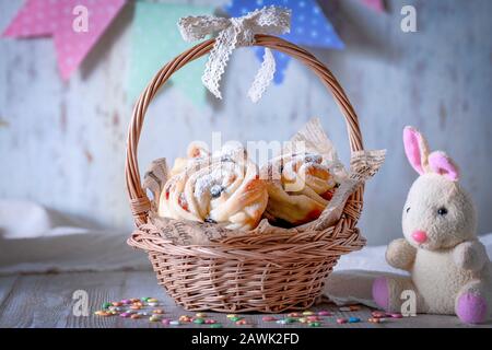 Easter bunny next to a basket filled with pastries. Kruffins with raisins and dried apricots, sprinkled with powdered sugar on top. Festive mood. Stock Photo