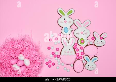 Top view of small chocolate balls in crispy sugar shell isolated on pink pastel background. Easter ginger glazed cookies in shape of bunnies, eggs in bright pink nest. Concept of easter holidays. Stock Photo