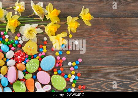 Crop of colorful ginger glazed cookies and chocolate balls and daffodils isolated on wooden background. Close up of homemade lovely delicious pastry in shape of easter animals, eggs and red flowers. Stock Photo