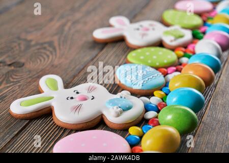Selective focus of big and small peanut chocolate balls in crispy sugar shell isolated on wooden background. Close up of easter ginger glazed cookies in shape of two bunnies and eggs in row. Stock Photo