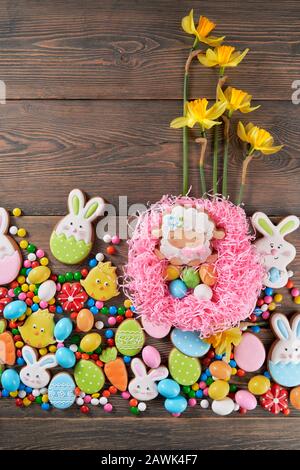Close up of peanut chocolate balls in crispy sugar shell and daffodils isolated on wooden background. Easter ginger glazed cookies in shape of animals, colorful eggs and sheep in pink nest. Stock Photo