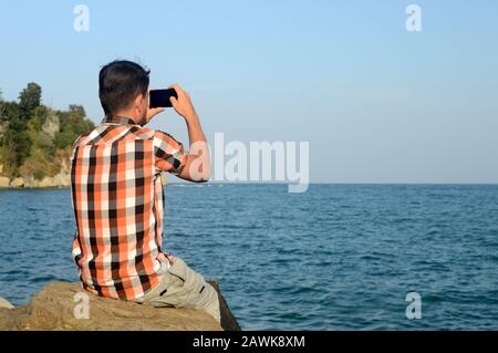 A young man, a tourist with smartphone, explores new places and shoot photos and makes a video of the summer seascape to share on social networks Stock Photo