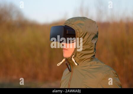 young soldier with virtual reality headset and in a green protective jacket with hood outdoors during a simulation of a combat situation Stock Photo