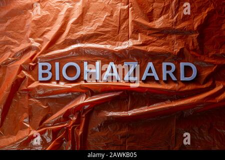 the word biohazard laid with silver letters on red crumpled plastic film background in flat lay composition at center with dramatic cold light Stock Photo