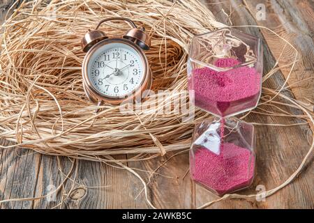 Pink hourglass and alarm clock on straw on wooden floor. Concept of Time Stock Photo