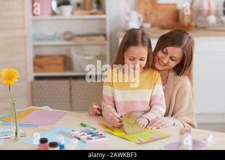 Warm-toned portrait of happy mother hugging daughter while drawing pictures at wooden kitchen table, copy space Stock Photo