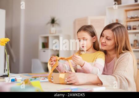 Side view portrait of happy mother hugging daughter while opening presents at wooden kitchen table, copy space Stock Photo