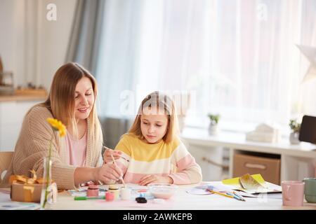 Warm-toned portrait of smiling mature mother painting pictures with cute little daughter while sitting at wooden table in cozy home interior, copy space Stock Photo