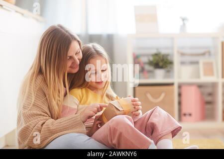 Warm-toned portrait of mother and daughter holding present while sitting on floor in cozy childrens room interior, copy space Stock Photo