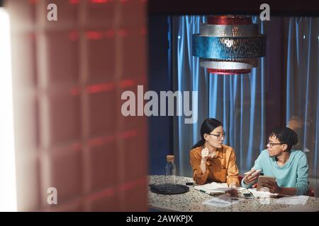 Wide angle portrait of two ethnic business people discussing project while working late in dark office, copy space Stock Photo