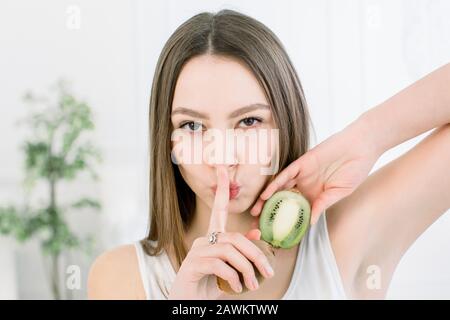 Beauty portrait of a pretty smiling woman with healthy skin showing silence gesture with finger over her lips and holding kiwi slices, isolated over Stock Photo