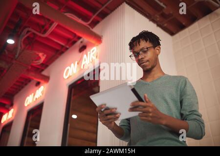 Low angle portrait of young African-American businessman using tablet while standing by ON AIR sign at radio, copy space Stock Photo