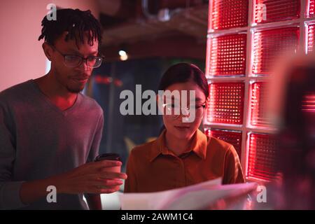 Multi-ethnic group of business people discussing documents while standing outdoors in dim red lighting, copy space Stock Photo