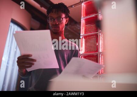 Low angle portrait of contemporary African man reading documents while standing outdoors in dim red lighting, copy space Stock Photo