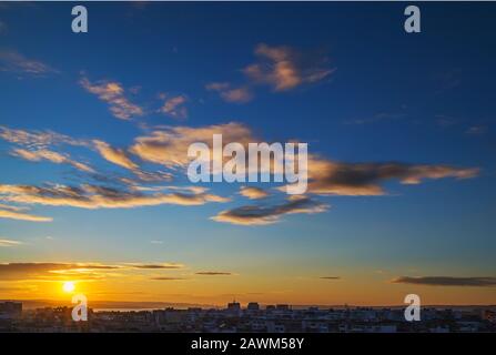 Scenic cityscape with golden evening sun goes down over the horizon. Gold colored sunset clouds in a clear blue sky. Beauty in nature. Stock Photo