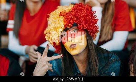 Woman sitting in fan zone with a wig and face painted in german flag colors gesturing victory sign. German soccer supporter in stadium looking at came Stock Photo