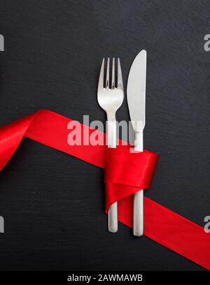 Valentine day dinner table setting. Fork and knife cutlery tied with passion red color ribbon on black background, top view. Stock Photo