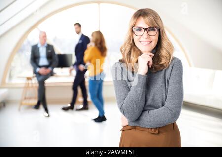 Portrait shot of executive middle aged businesswoman standing in the office while business people consulting in the background. Stock Photo