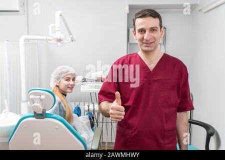 portrait of a happy dentist doctor showing thumbs up on the background of his office with a patient in a chair Stock Photo