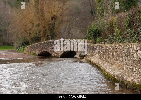 Ancient horse bridge in Dunster Somerset. Looking along the river with path to the right. Flowing river passing under the stone structure. Stock Photo