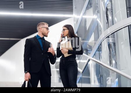 Business couple on escalator talking and smiling. Man wearing suit and glasses with a bag talking with a woman holding tablet computer Stock Photo