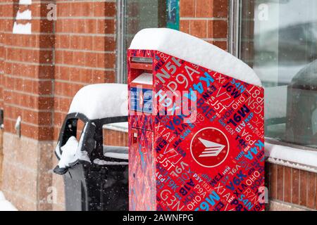 A Canada Post street letter box stands outside shops in an outdoor shopping plaza during a winter snowfall, with a layer of snow on the mailbox. Stock Photo