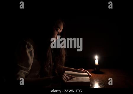 A young girl reading a book at night with candle light. She is wearing an old white dress. Dark background. Side view. Stock Photo