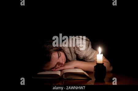 A young girl sleeping on an old book at night with candle light. The girl is wearing an old white dress. Dark background. Front view. Stock Photo