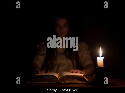 A young girl is holding a book at night. She is wearing an old white dress. The focus is on the book. The girl is looking into the camera. Stock Photo