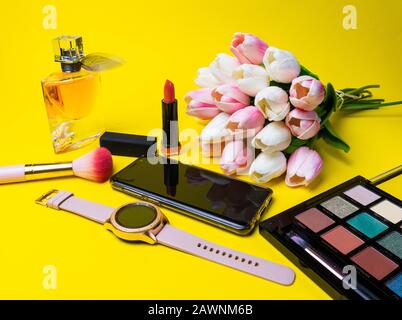Makeup products perfume, brush, flowers clock, mobile phone multi colored palette on yellow background Stock Photo