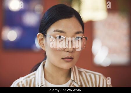Head and shoulders portrait of young Asian woman wearing glasses looking at camera while posing against red wall in cafe, copy space Stock Photo