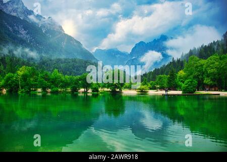 Mountain lake landscape, with lush green pine forrest,  misty clouds and sun peeking from behind a peak Stock Photo