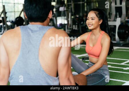 Face of Young Asian mixed sporty woman chatting with a muscuar man sitting on the floor at fitness gym. Healthy couple concept Stock Photo