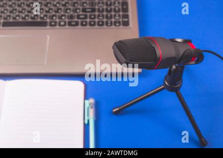 A microphone for recording sound and podcasts next to a laptop and notebook on a blue background. Desktop of blogger. Stock Photo