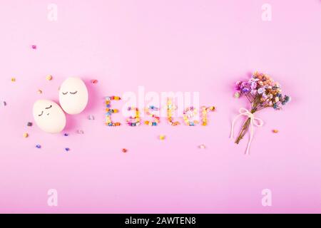 Easter creative layout with the word Easter made of flowers, a bouquet of small flowers and cute white eggs with closed eyes on the lilac. Stock Photo