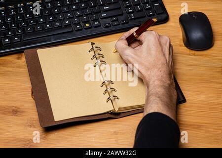 Desktop workspace with keyboard and mouse partially visible.  Vintage style notebook with a finely crafted wooden pen being used by a caucasion male h Stock Photo