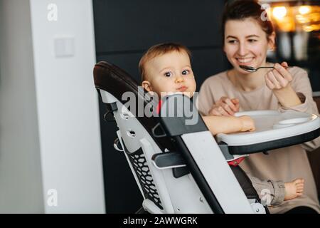 Child sitting on a high chair, getting breakfast from his mother Stock Photo