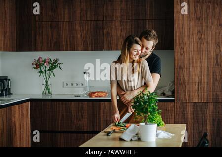 Man seeks attention, hugging his girlfriend while she's slicing vegetables Stock Photo