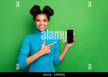 Photo of funny dark skin lady hold telephone smart phone new model sale price directing finger touch screen wear blue turtleneck jumper isolated green Stock Photo