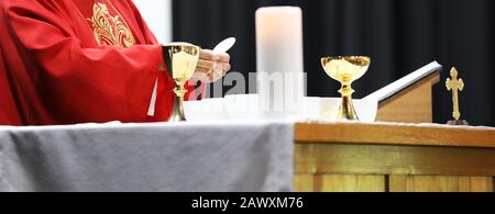 A Catholic Priest about to break the bread host while celebrating blessed Holy Communion at Mass. Wearing a red gown vestment and surrounded by chalic Stock Photo