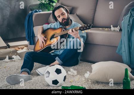 Photo of hipster guy with long beard sitting carpet near sofa holding guitar don't mind chaos after stag party messy dirty flat singing songs careless Stock Photo