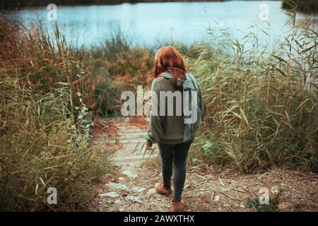 Young redhead woman walking to the lake shore with headphones wearing a jersey and black jeans Stock Photo
