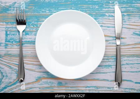 Empty white plate with silver fork and knife on blue wooden table. Dinner place setting. Top view Stock Photo