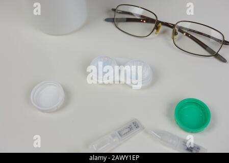 Flat lay of various eye care products on white counter. Contact lens case opened with glasses, droppers and solution surrounding Stock Photo
