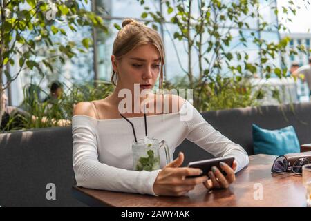 Upset young student watching broadcasting online on modern mobile phone. Blond woman blogger with cocktail is reading incoming sms message on Stock Photo