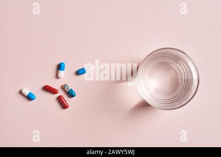 Various capsule pills and glass of water on pale pink surface, horizontal from above flat lay shot Stock Photo