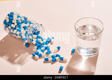 White And blue capsules spilled out of fallen bottle and glass of water on pale pink table surface, horizontal shot Stock Photo