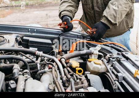 Mechanic engineer charging car battery with electricity using jumper cables outdoors. Red and black Jumper cables in male hands of car mechanic. Man Stock Photo