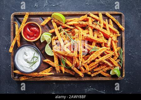 Sweet Potato Fries with sauce and ketchup on a rude wooden board on a concrete table with lime and bouquet of aromatic herbs, horizontal view from abo Stock Photo